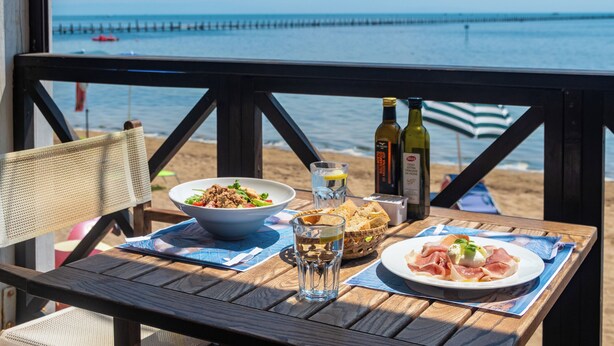 Una mesa de madera con un tazón de ensalada, un vaso de agua, una botella de vino y un plato de comida en una terraza con vista al mar.