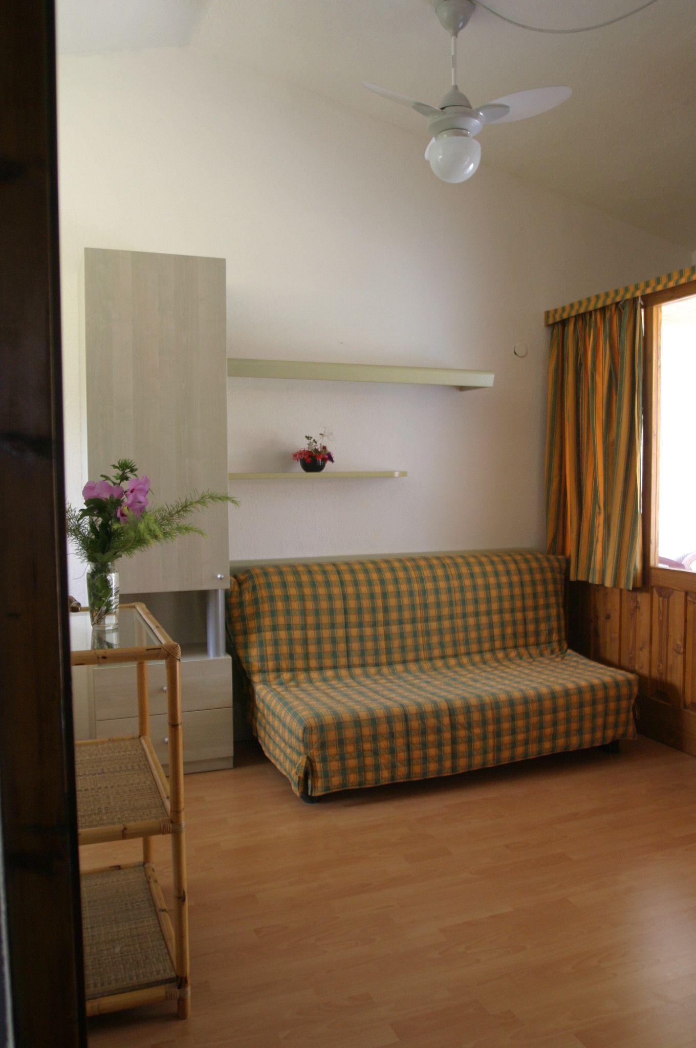 Una habitación con piso de madera, un sofá de cuadros, un ventilador de techo, una mesita con un jarrón de flores y una ventana con cortinas.