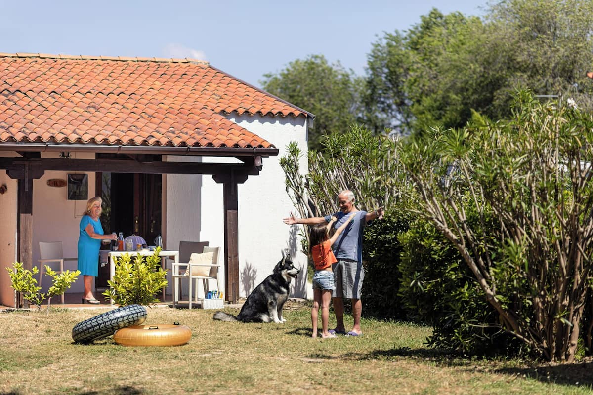 A house with a tiled roof and a covered porch. There is a dog sitting on the grass near the house.