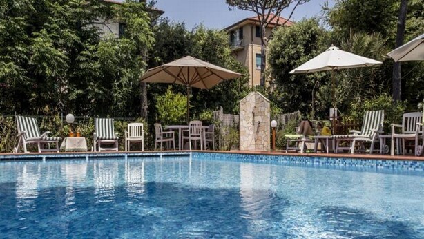 Ein Swimmingpool mit Liegestühlen und Sonnenschirmen, umgeben von Grünflächen und Gebäuden.