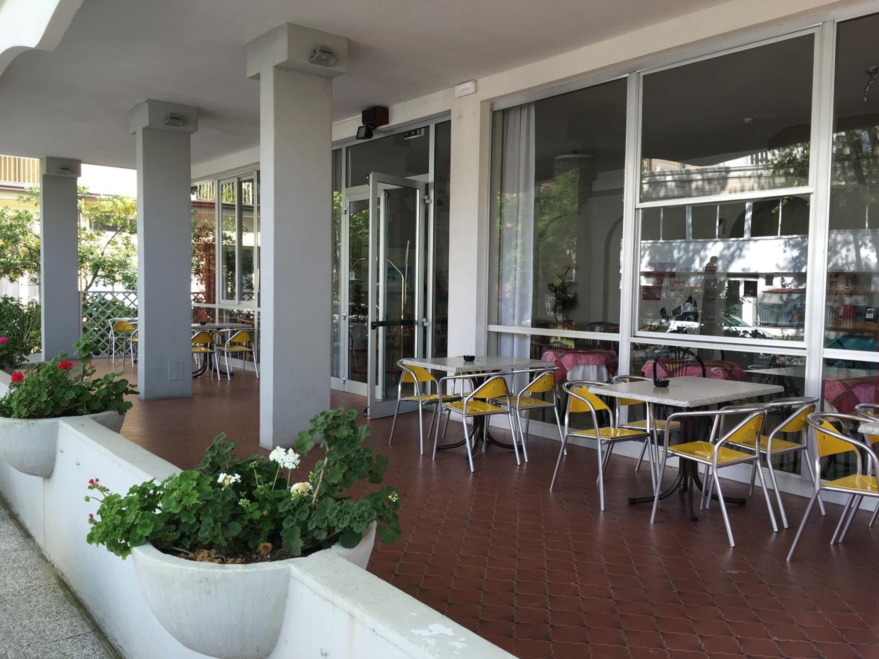 Ein Café im Freien mit Tischen, Stühlen, großen Fenstern und Topfpflanzen.