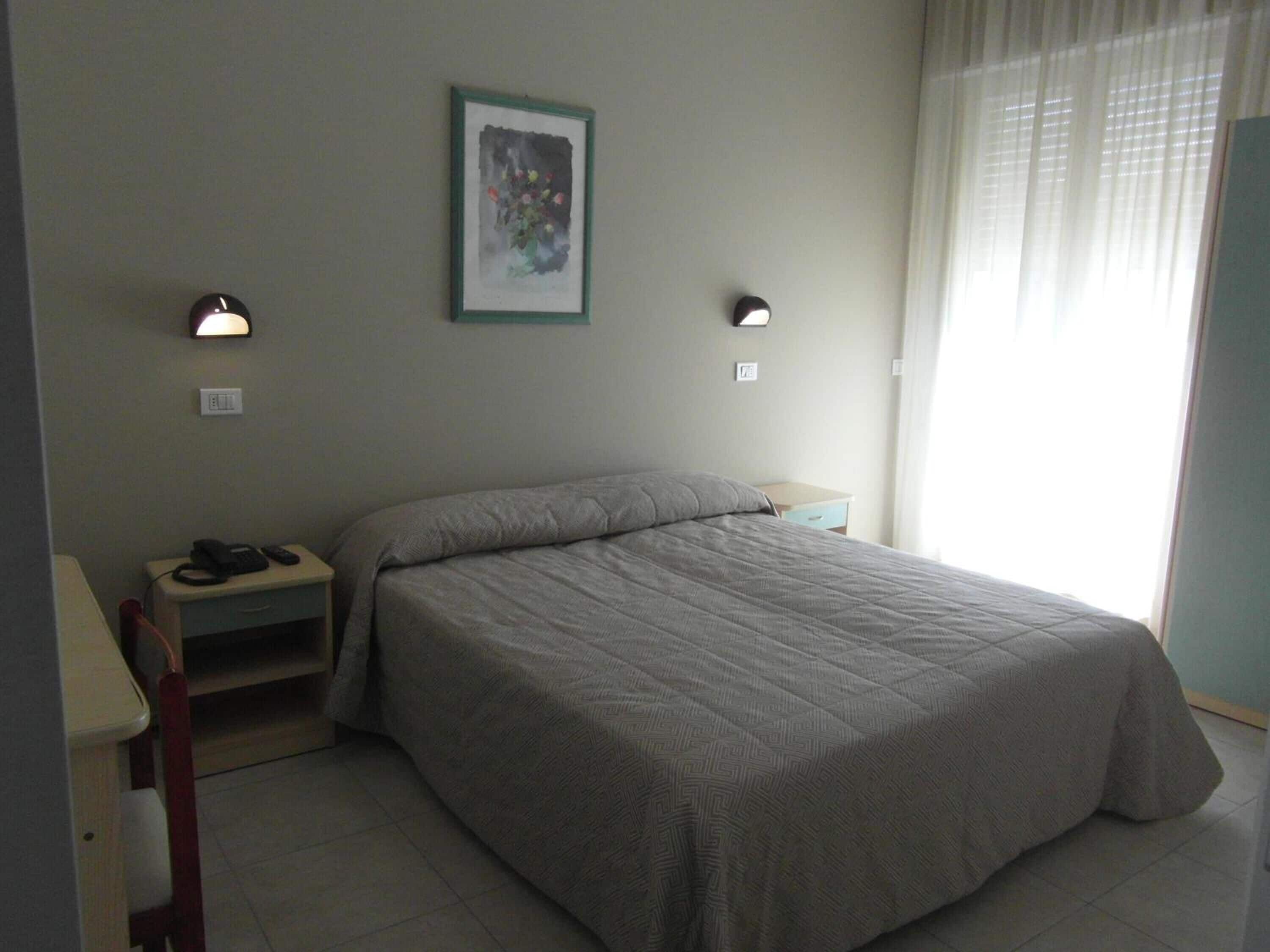 Une chambre à coucher comprenant un lit, des tables de chevet, une commode, une lampe fixée au mur et une fenêtre avec des rideaux.