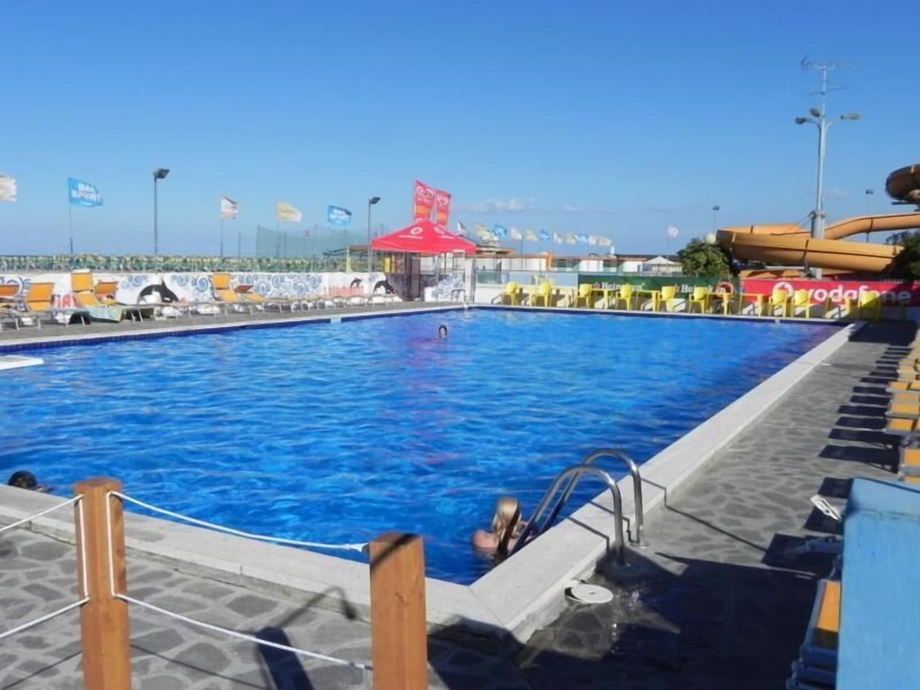 Une piscine avec une terrasse carrelée, un poste de surveillance pour les sauveteurs et un toboggan aquatique en arrière-plan.