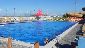 Une piscine avec une terrasse carrelée, un poste de surveillance pour les sauveteurs et un toboggan aquatique en arrière-plan.
