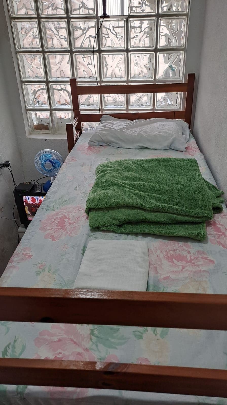 A small, single-bed room with a wooden frame, a floral-patterned bedspread, a green towel, and a window with a view of trees.