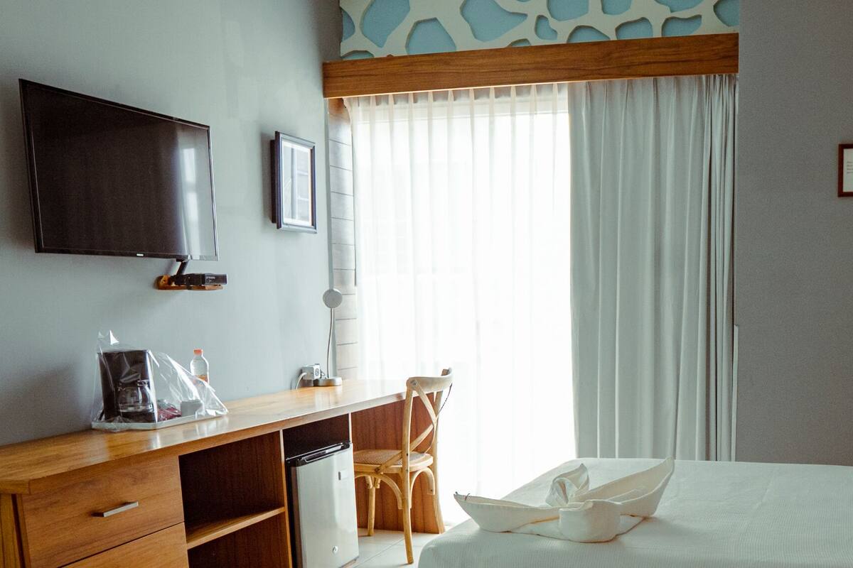 A hotel room with a bed, a desk with a chair, a television, a window with curtains, and a framed certificate on the wall.