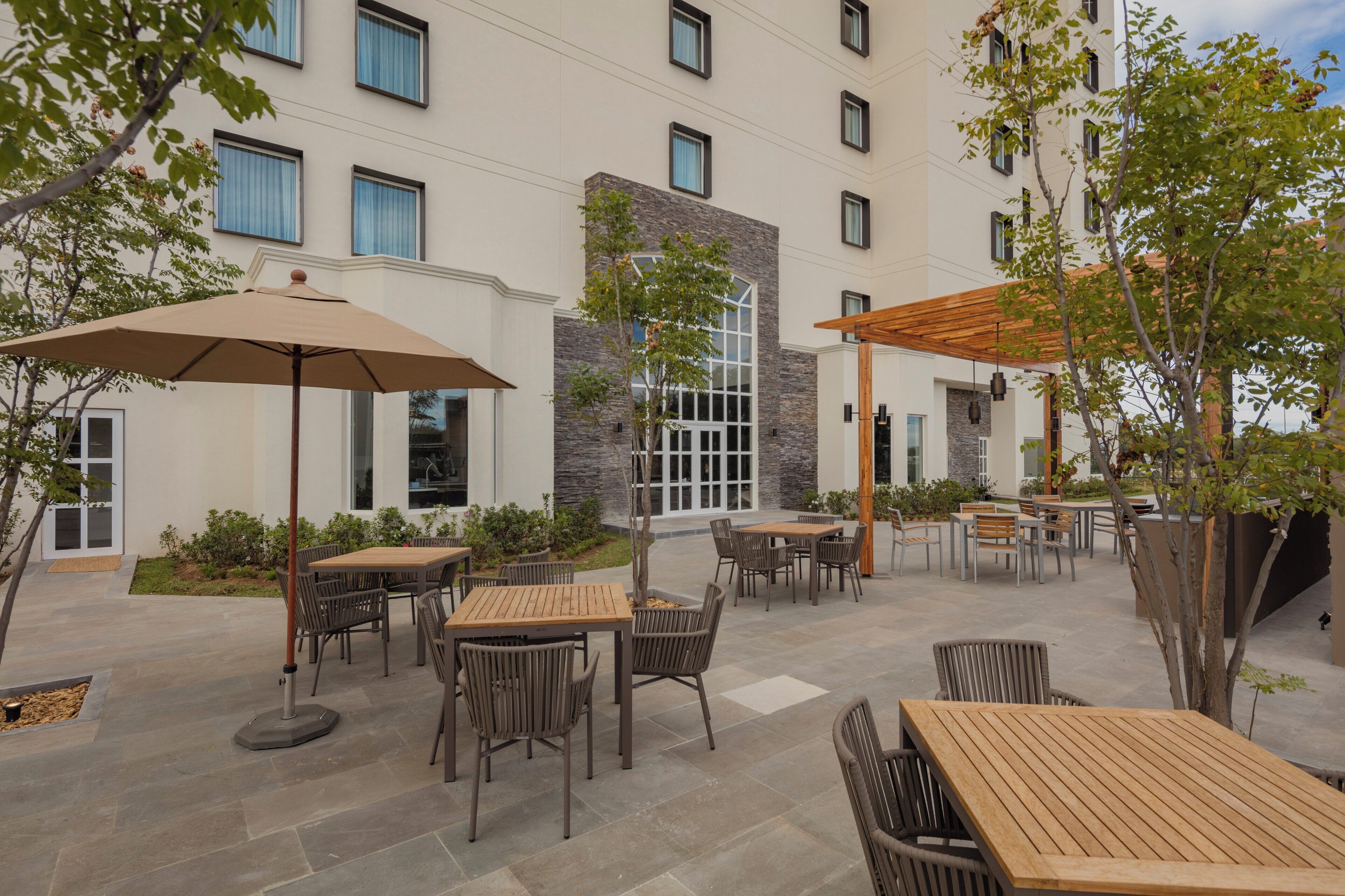 An outdoor patio area with multiple tables and chairs, a pergola, and a building with a stone facade and large windows.
