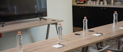 Un salón de conferencias con una larga mesa, sillas, un televisor y botellas de agua.