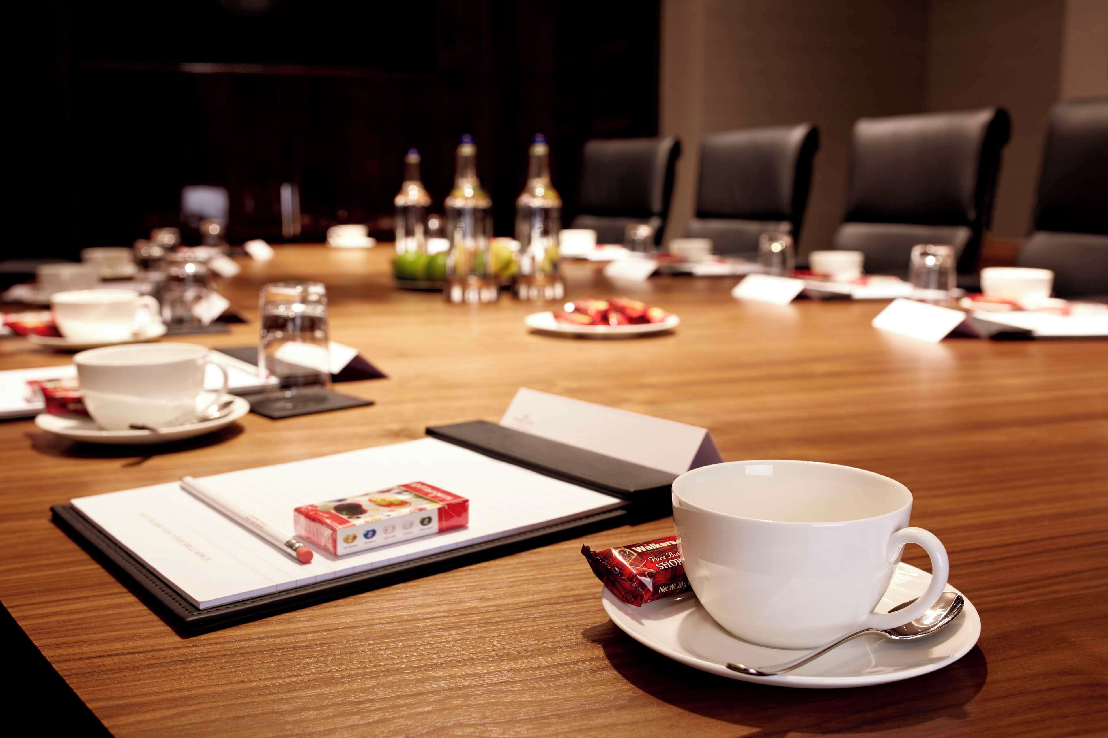 A conference table with a white cup, saucer, and spoon, a notepad, a box of tissues, and a glass of water.