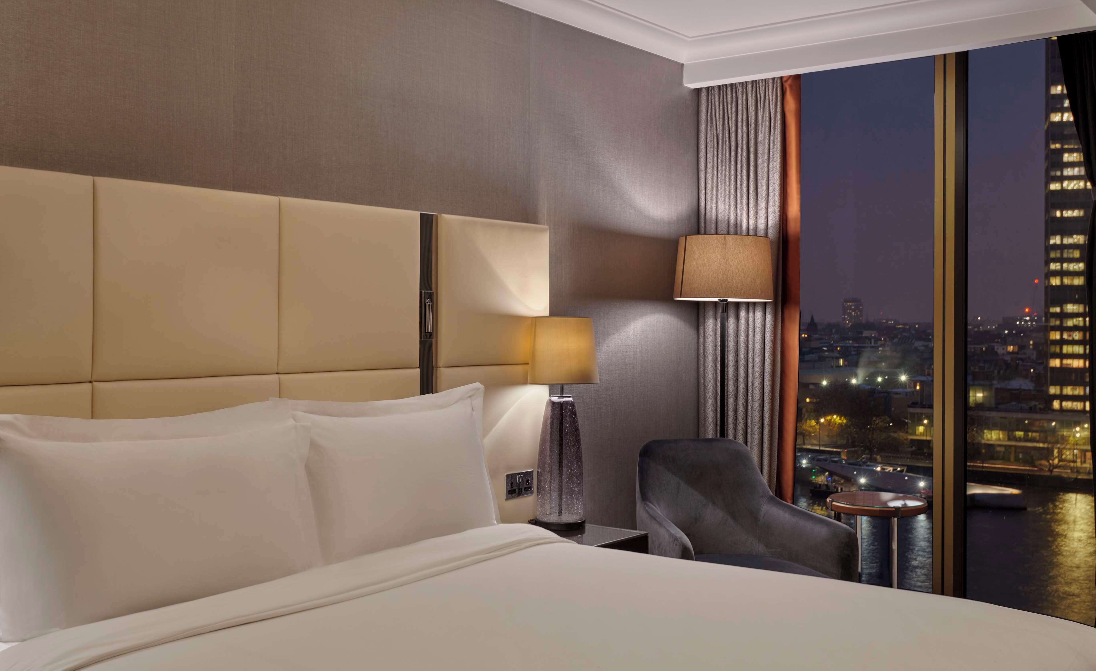 Una habitación de hotel con una cama grande, una mesita de noche, una lámpara, una silla y vistas a un paisaje urbano nocturno.