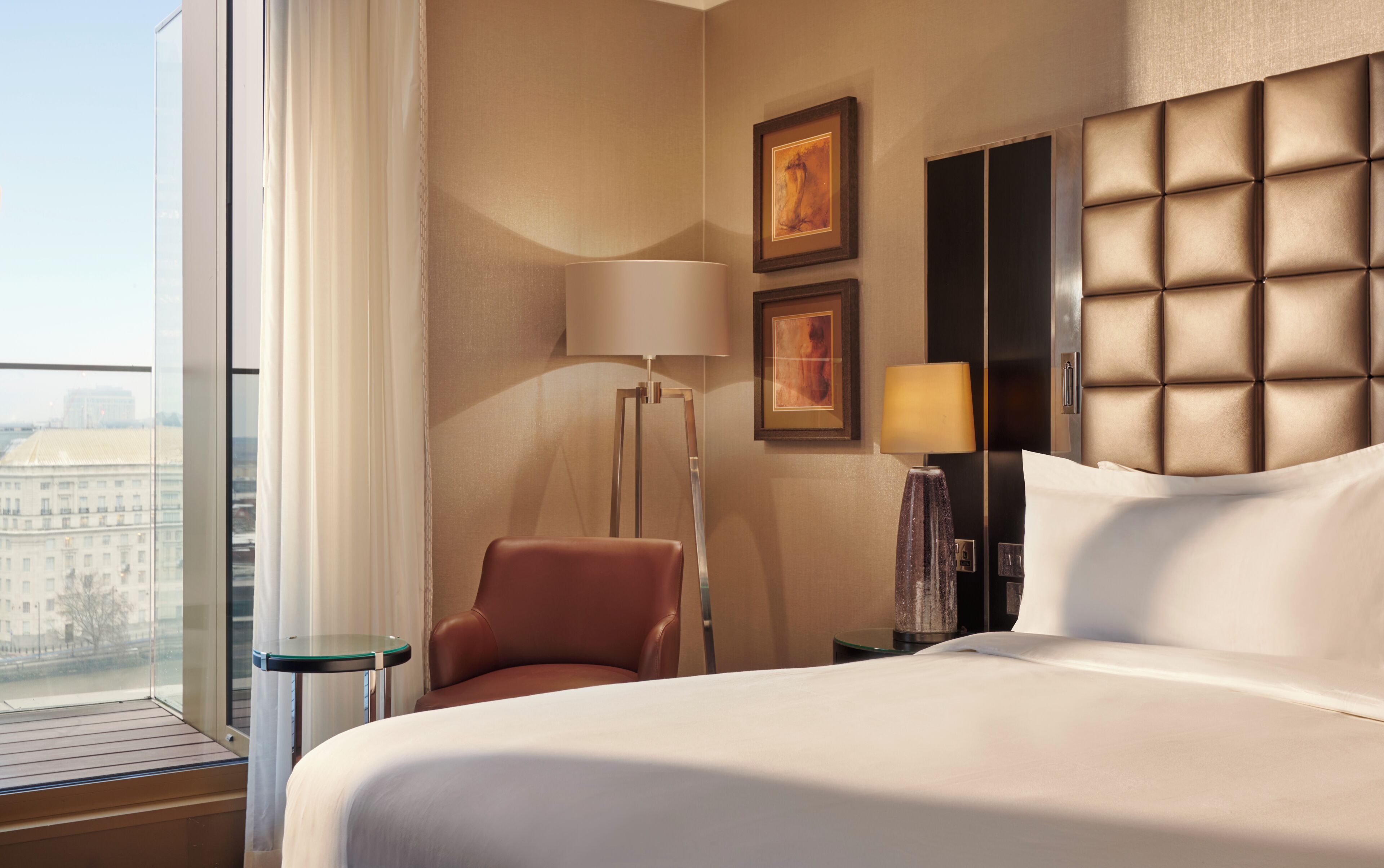 A hotel room with a bed, a chair, a floor lamp, a bedside table, and a large window.