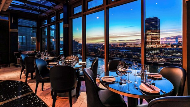 Een restaurant met grote ramen dat uitzicht biedt op de stad in de schemering. Tafels zijn gedekt met wijnglazen en borden.