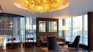 Een moderne hotel lobby met een grote kroonluchter, een zithoek en grote ramen met dunne gordijnen.