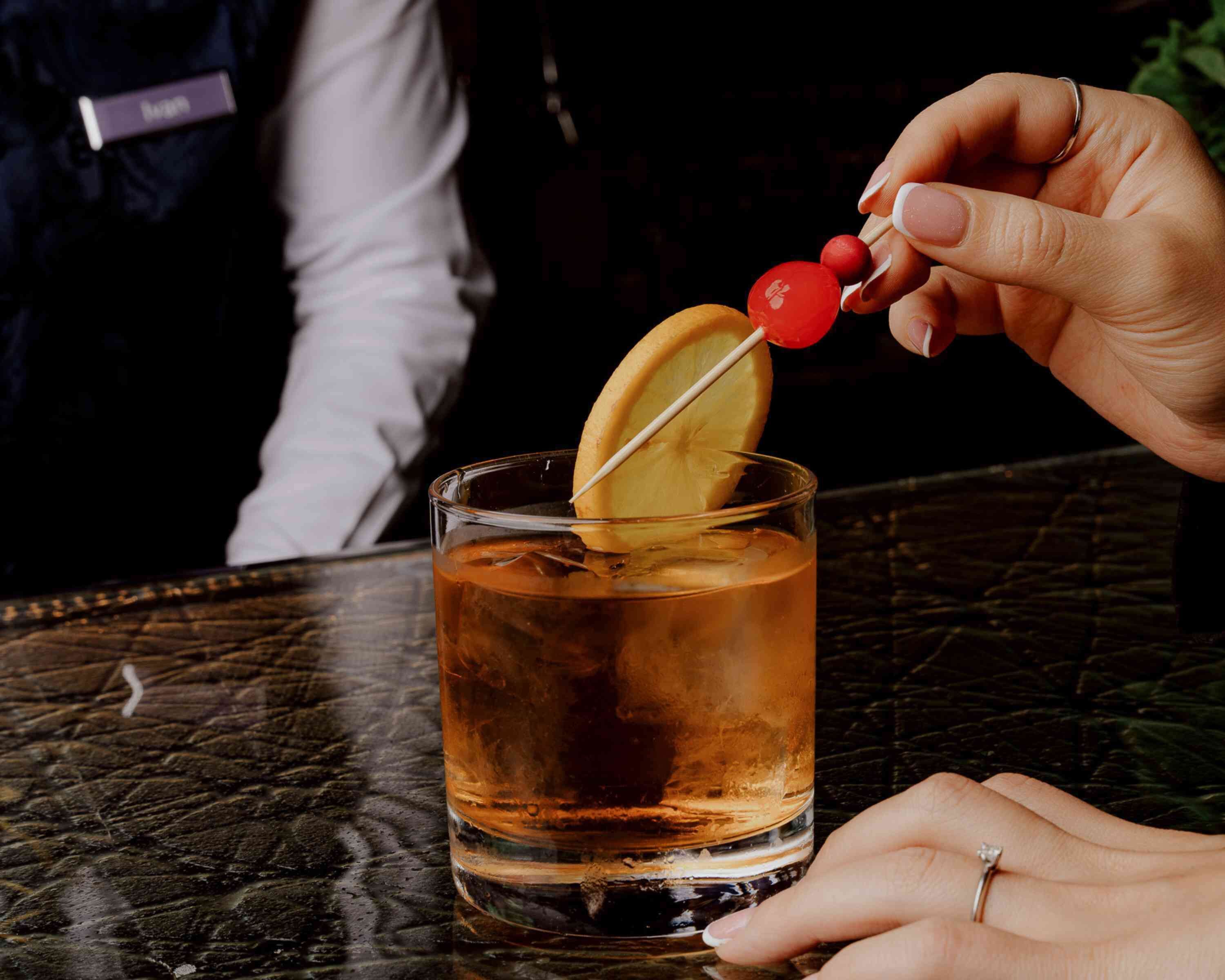 A person holding a cocktail with a slice of lemon and a cherry on a skewer, with a glass of amber-colored drink in the foreground.
