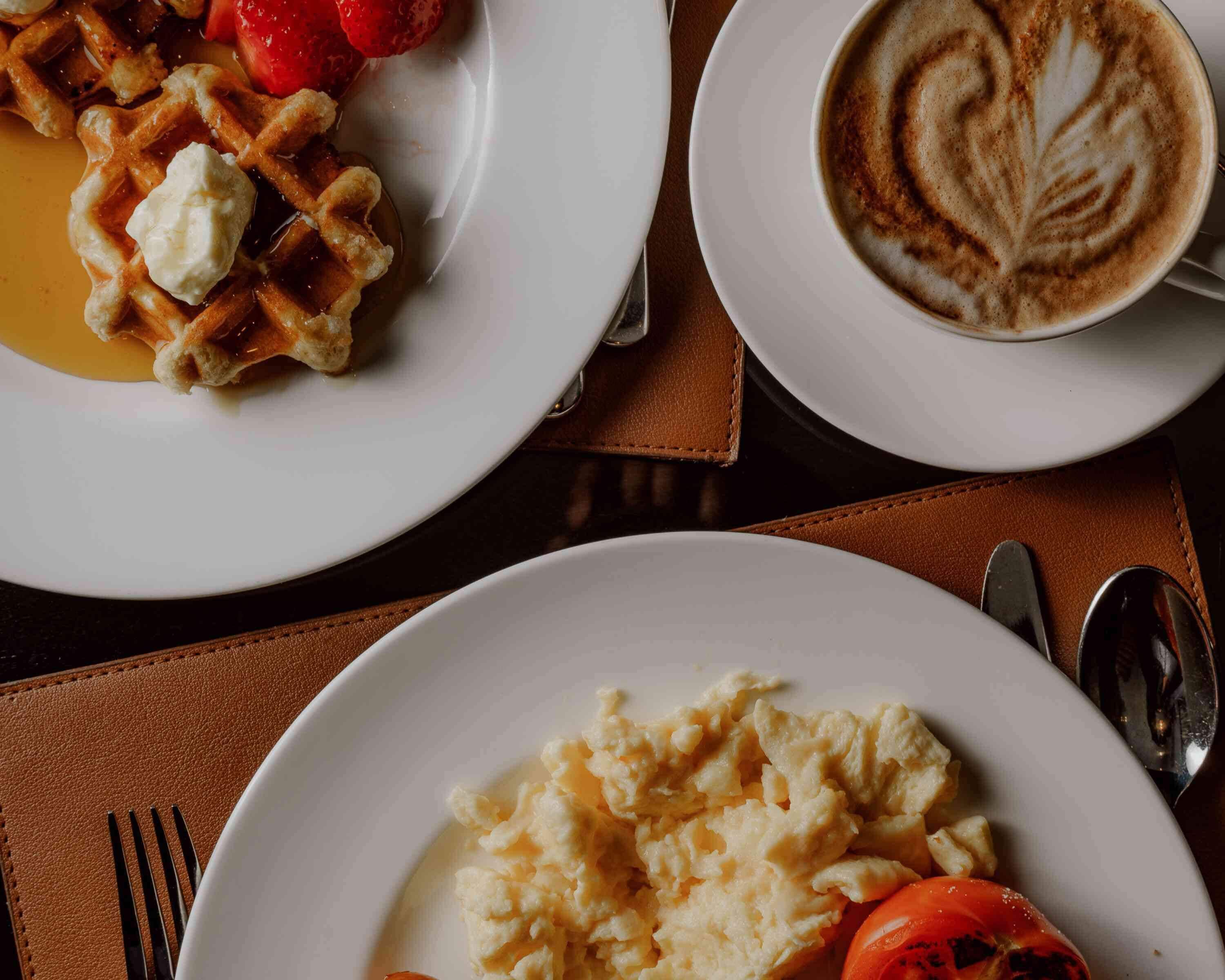 A breakfast setting with waffles, scrambled eggs, a slice of tomato, and a cup of coffee with latte art.
