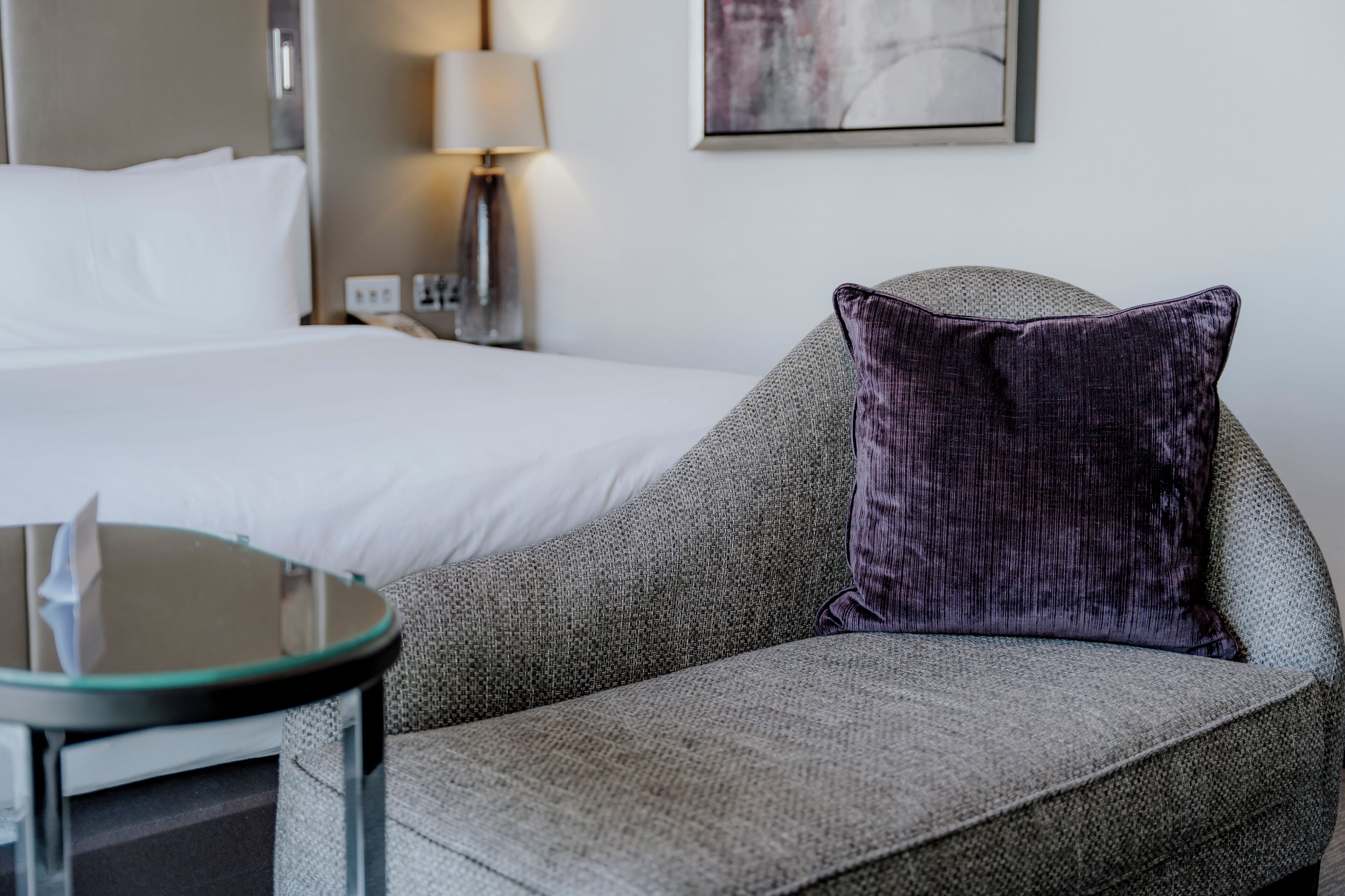 A hotel room with a bed, a grey sofa with a purple pillow, a small round table, and a lamp.