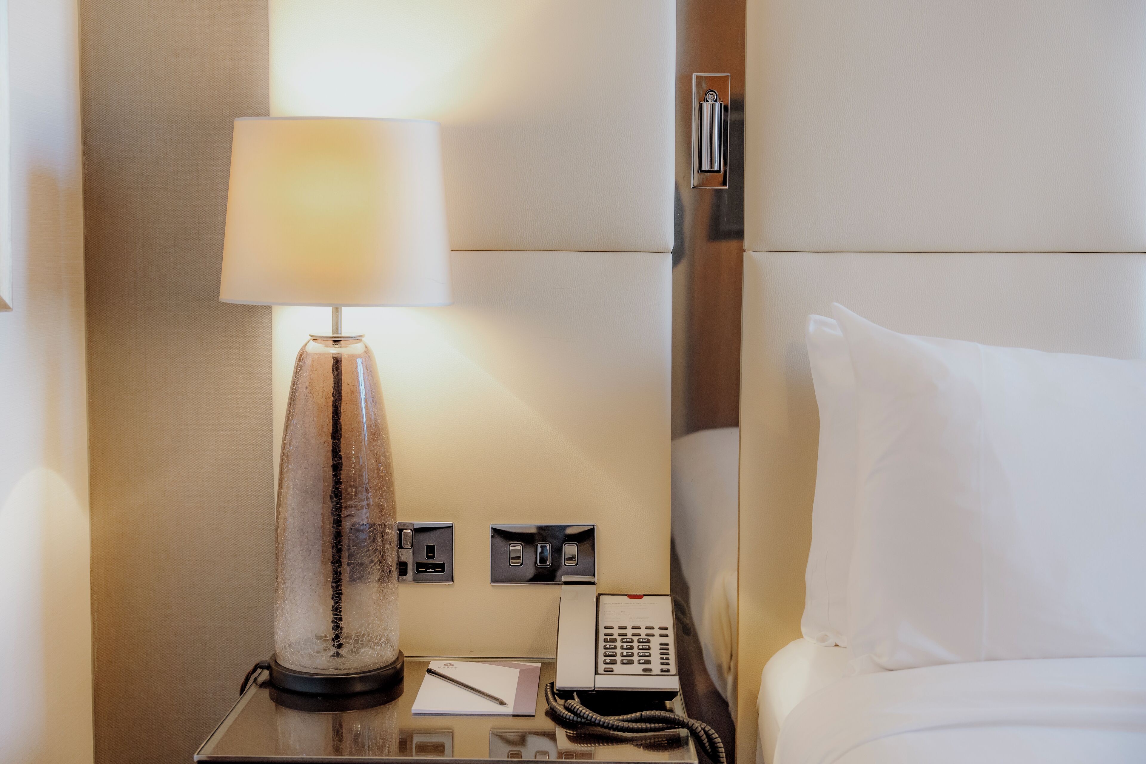 A hotel room with a bedside lamp, a telephone, and a bedside table with a notepad and pen.