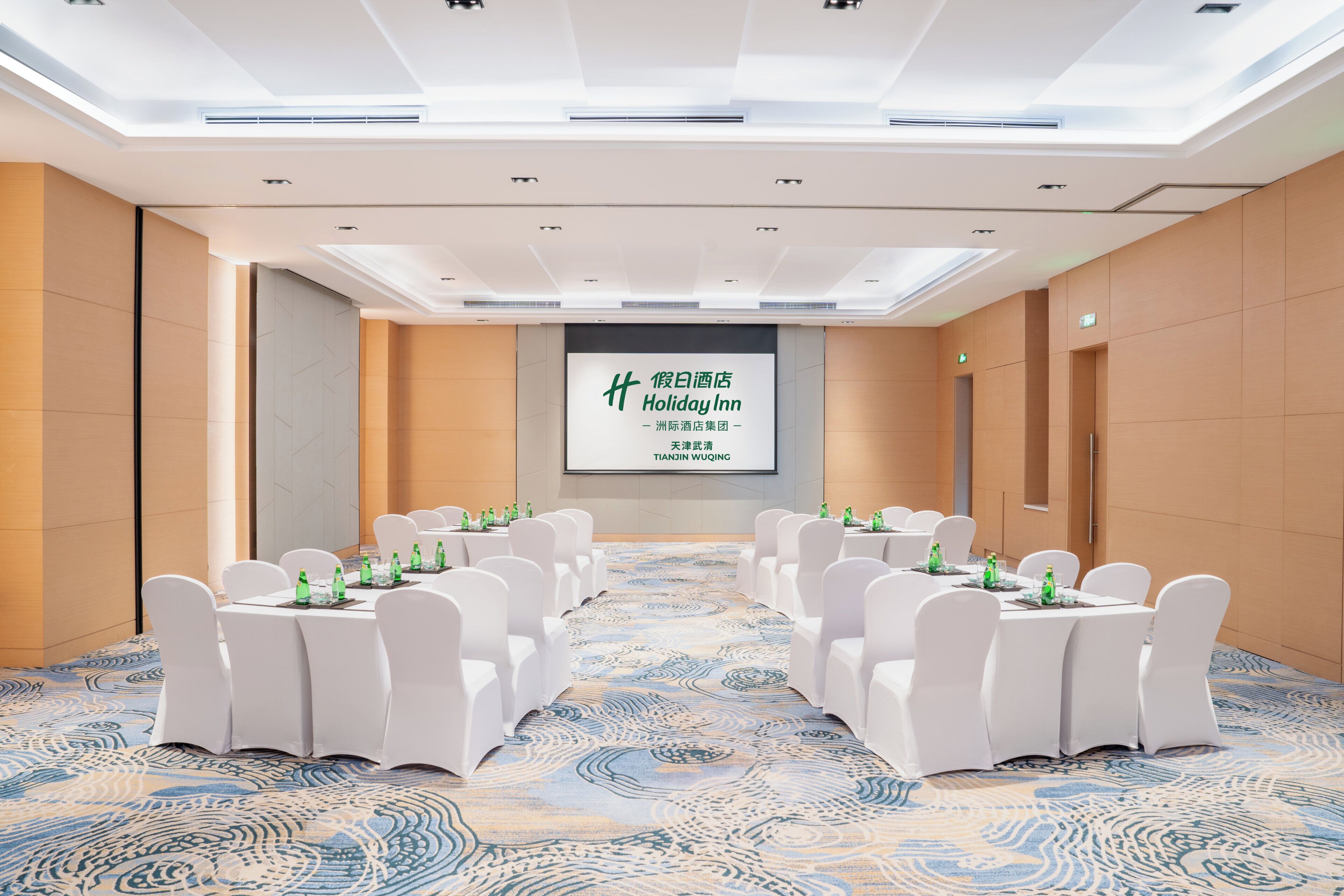 Un salón de conferencias con una pantalla grande mostrando la marca 'Holiday Inn', sillas blancas y mesas dispuestas para una reunión.