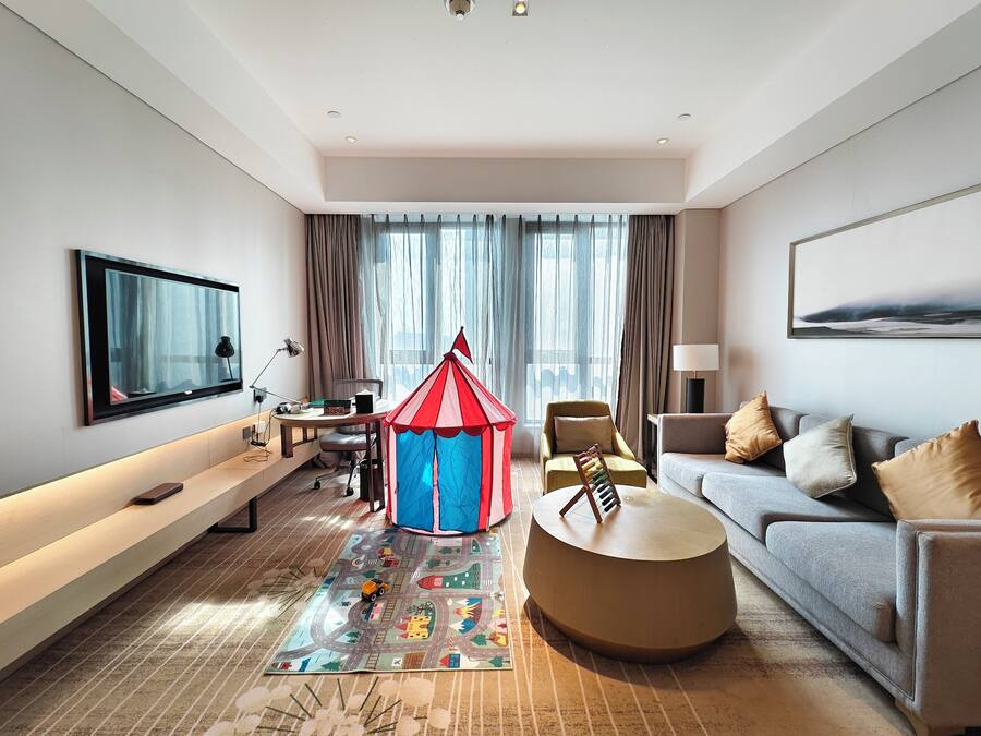 A modern hotel room with a flat-screen TV, a comfortable sofa, a small round coffee table, a children's play tent, and a large window with curtains.