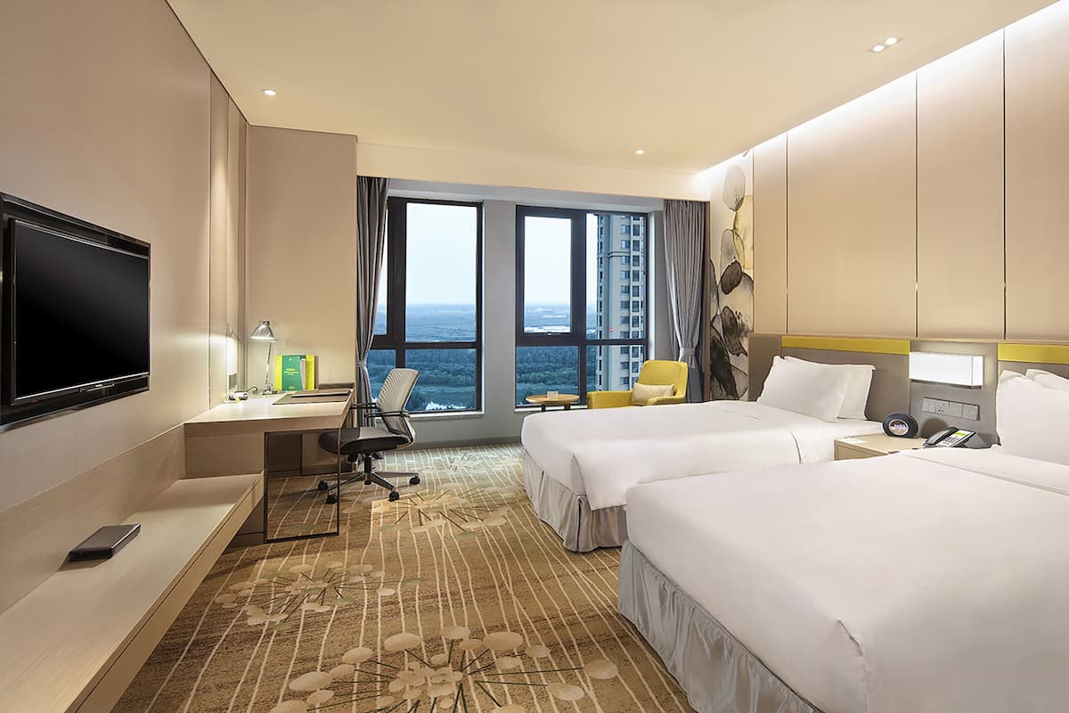 A hotel room with two beds, a desk with a chair, a TV, and a large window offering a view of the city.