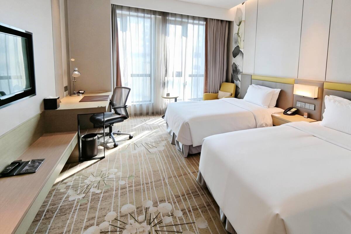 A hotel room with two beds, a desk, a chair, a TV, and a large window with curtains.