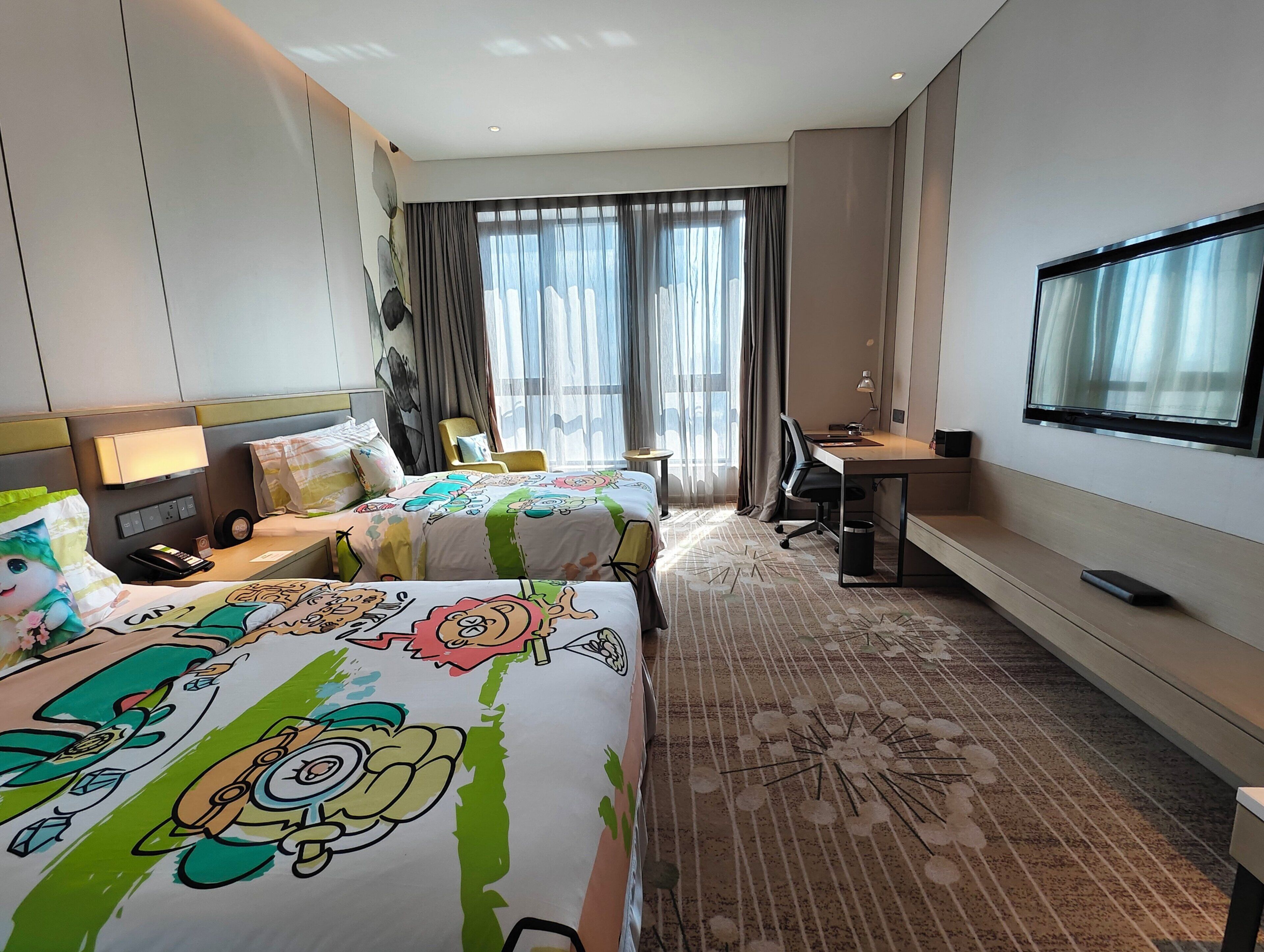 Habitación de hotel con dos camas, un escritorio, una silla, un televisor y un amplio ventanal con cortinas.