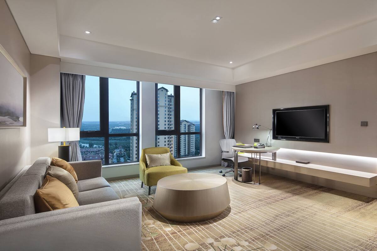 A modern hotel room with a large window, a flat-screen TV, a sofa, a desk, and a bed.