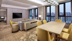 A modern hotel room with a large dining table, yellow chairs, a sofa, a TV, and large windows offering city views.