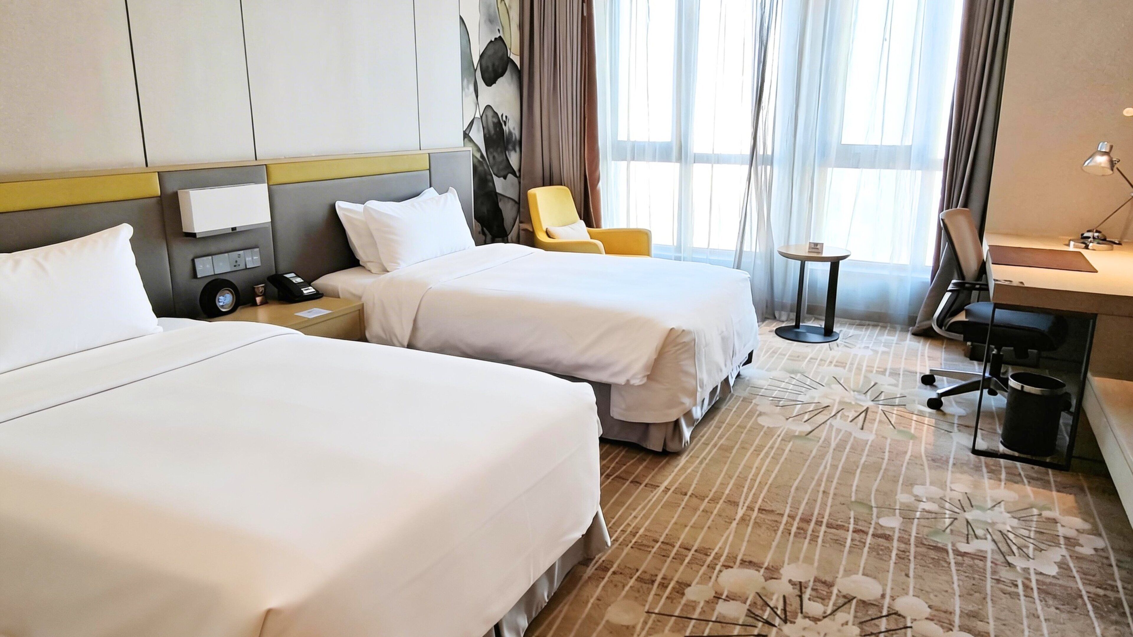 A hotel room with two beds, a desk, a chair, a lamp, and a window with curtains.