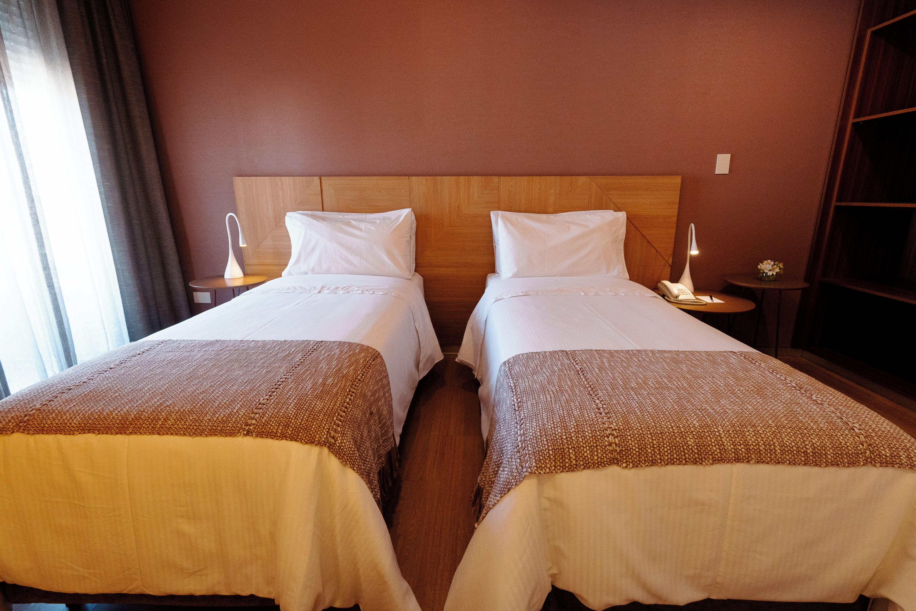 A hotel room with two single beds, wooden headboards, bedside tables with lamps, and a large window with curtains.