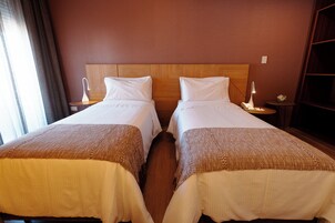 Habitación de hotel con dos camas individuales, cabeceros de madera, mesitas de noche con lámparas y un amplio ventanal con cortinas.