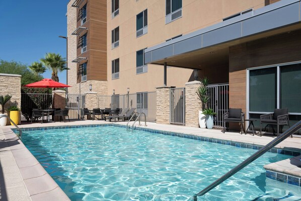 A swimming pool with lounge chairs and umbrellas, surrounded by a stone wall and a building with multiple windows.