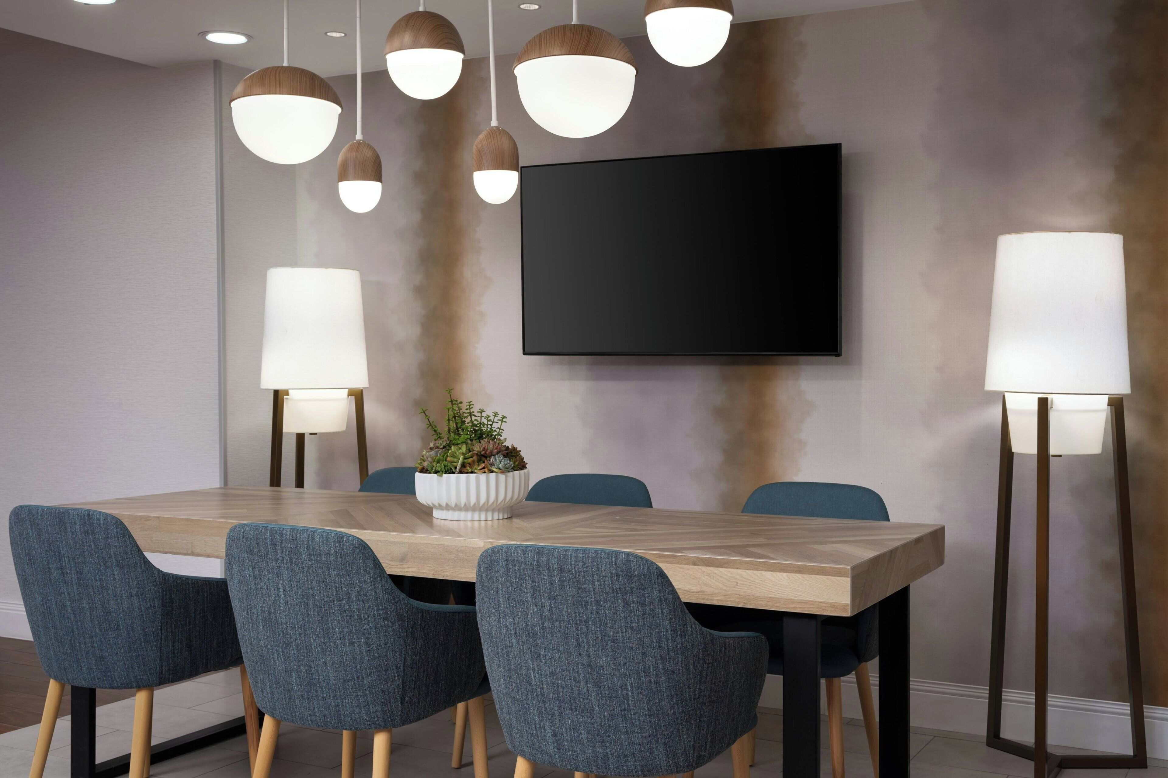 A modern dining area with a wooden table, six upholstered chairs, pendant lights, a large flat-screen TV, and floor lamps.