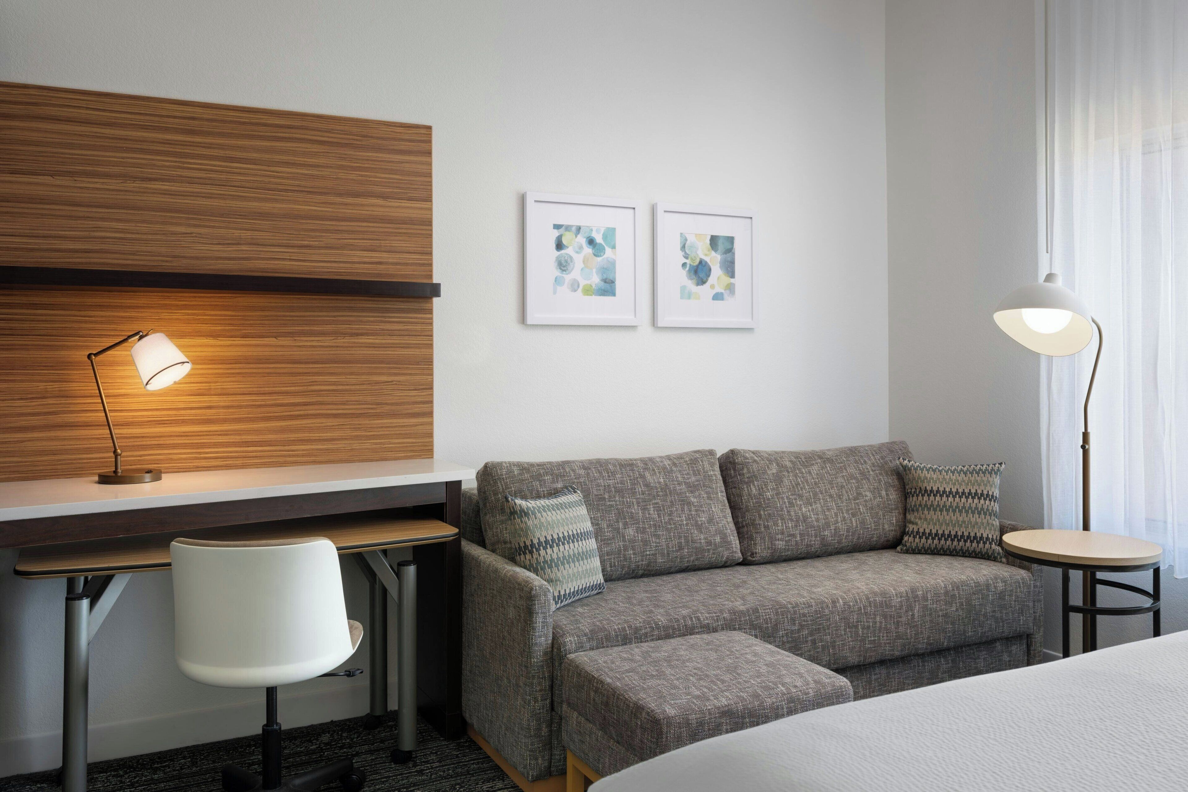 A modern hotel room with a grey sofa, a small desk with a lamp, a chair, and two framed artworks on the wall.