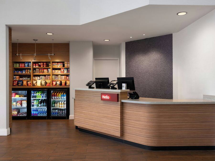 A reception area with a wooden reception desk, a 'Hello' sign, and a wall-mounted screen. There are refrigerated beverage dispensers in the background.