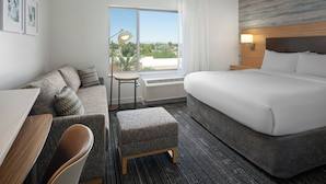 Habitación de hotel con cama, escritorio, silla, reposapiés, ventana con vistas y lámpara de pared.