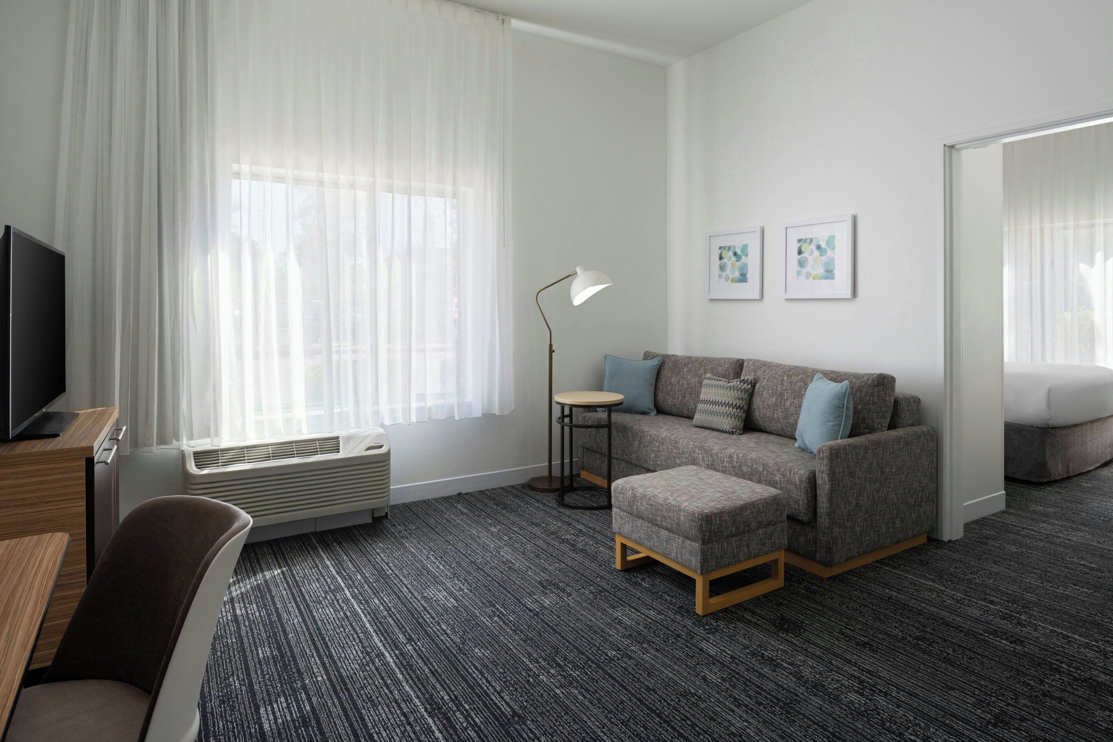 Ein modernes Hotelzimmer mit grauem Sofa, einem kleinen runden Tisch, einem Fernseher und einem großen Fenster mit leichtem Vorhang.