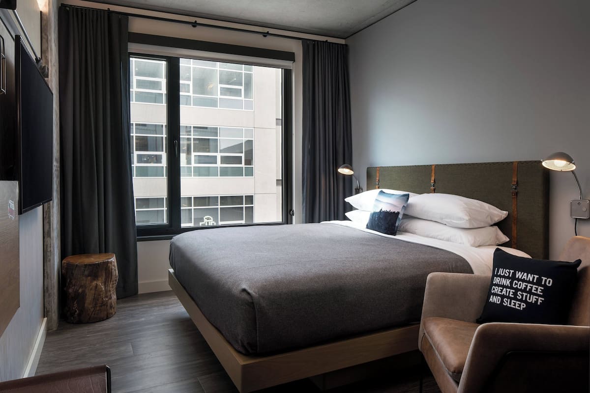 Quarto de hotel moderno com uma cama grande, uma mesinha de madeira, um banquinho e uma cadeira com almofada.