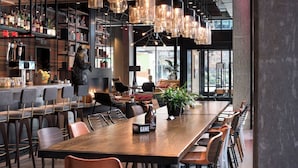 A modern bar area with a long wooden table, multiple chairs, and a variety of bottles and glasses. There are shelves with books and decorative items, and a large window providing a view outside. - MOXY Seattle Downtown (Seattle)