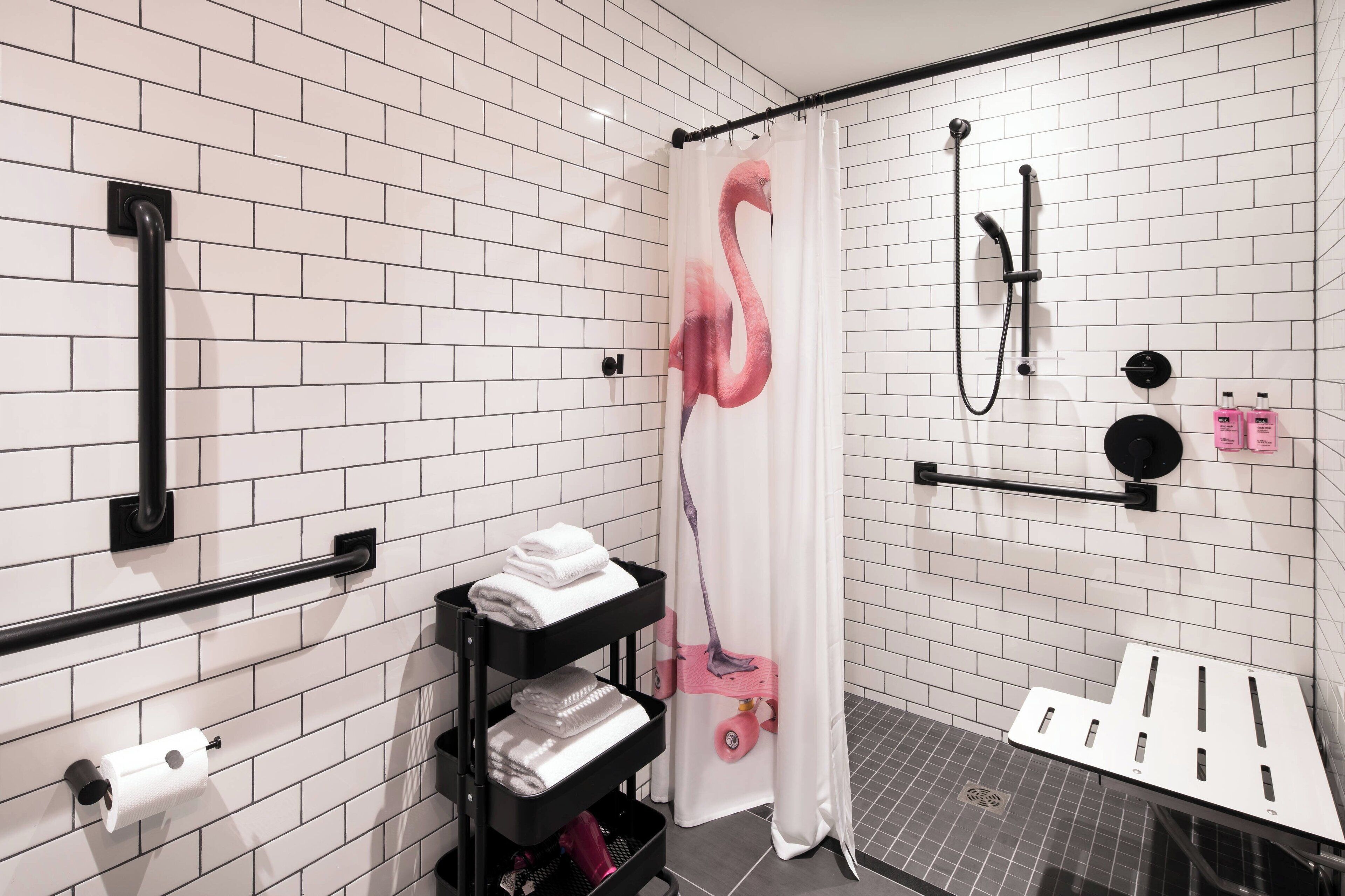 Un baño moderno con paredes de azulejos blancos, una ducha con cortina de flamencos rosados, un asidero negro y un estante con toallas.