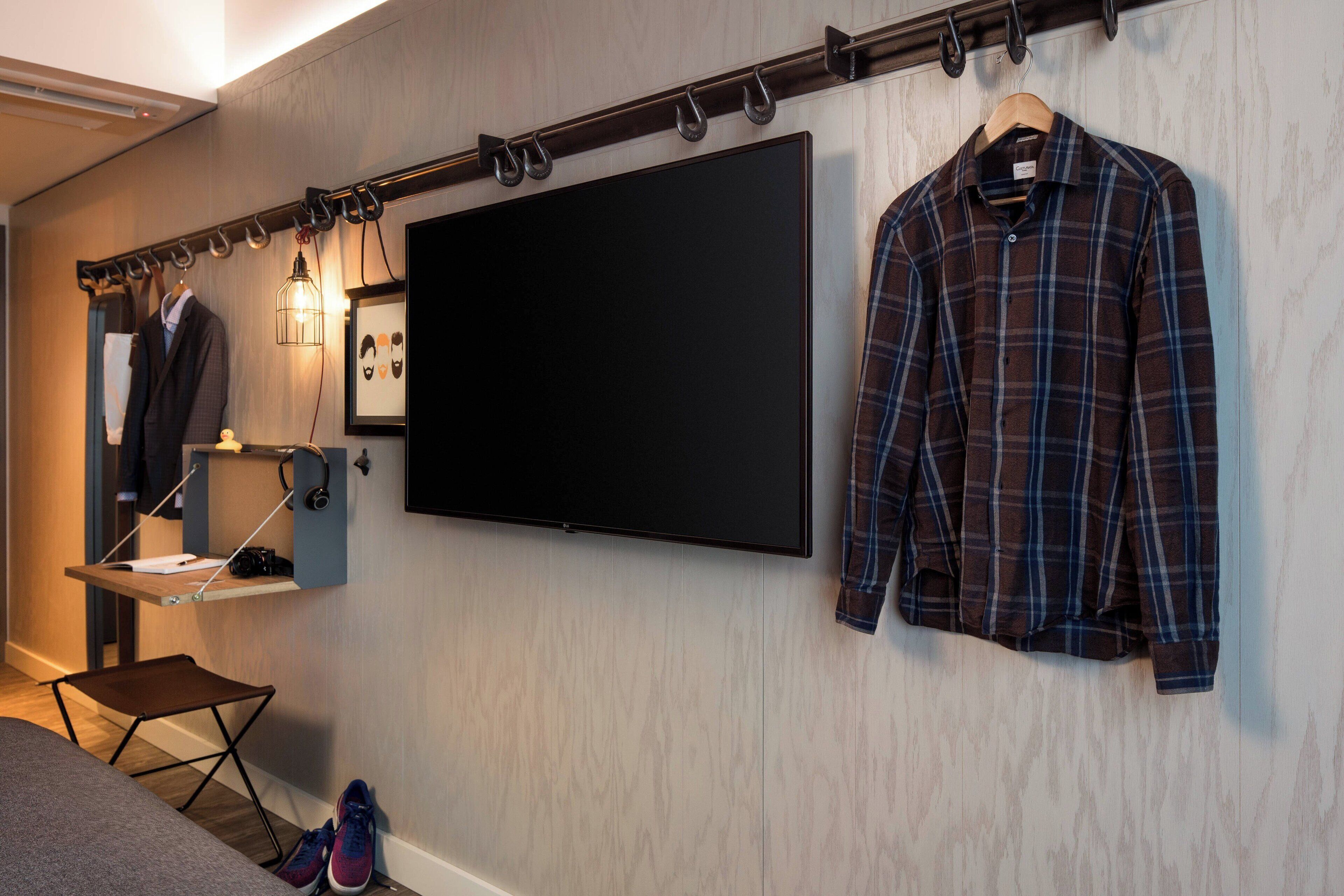 A room with a mounted TV, a coat rack with a plaid shirt, a mannequin with a suit, and a small table with a camera and papers.