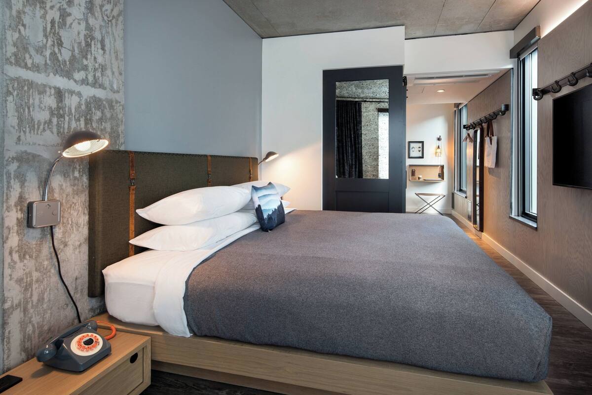 Quarto de hotel moderno com uma cama grande, luminárias de cabeceira, telefone e vista para o corredor e banheiro.