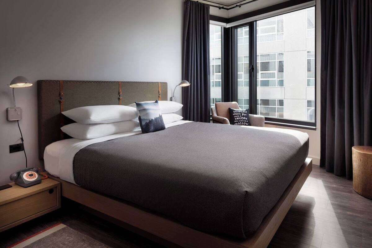 Quarto de hotel moderno com uma cama grande, mesa de cabeceira com luminária, uma cadeira e vista para a cidade através de amplas janelas.