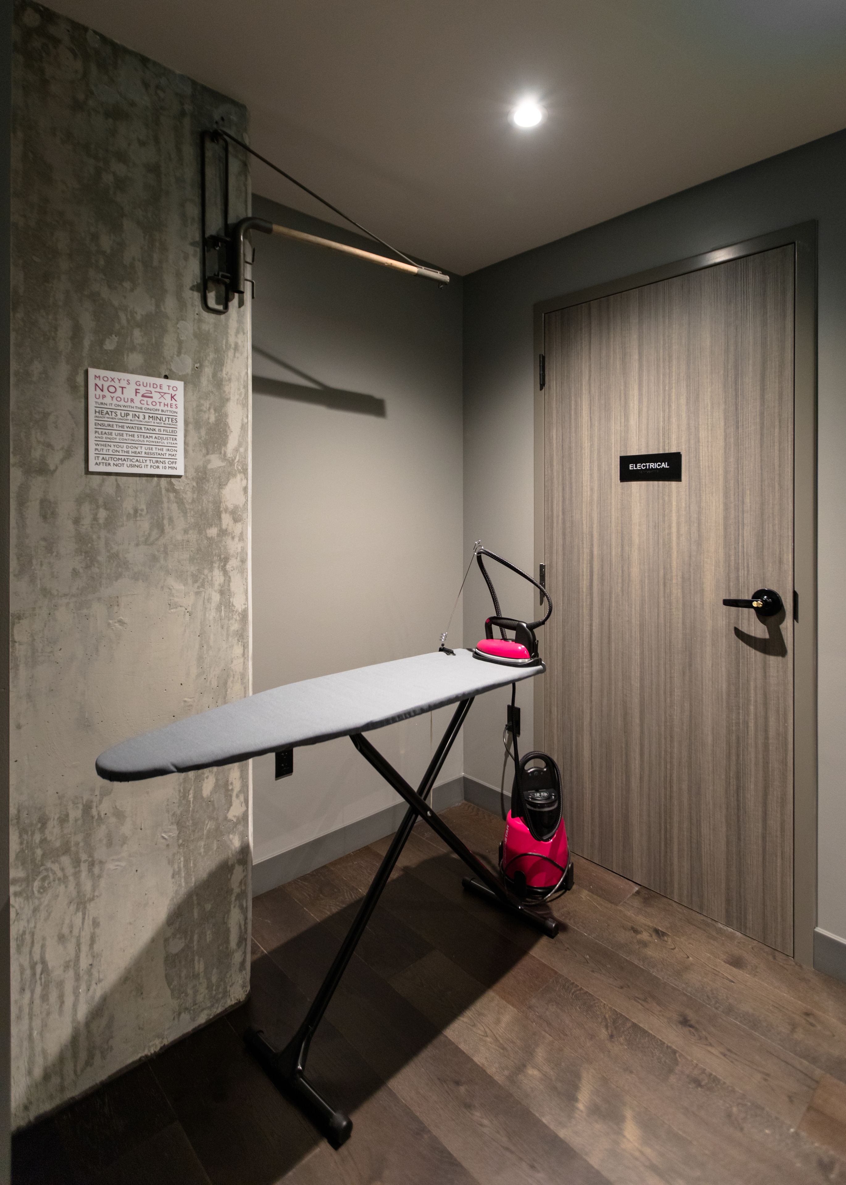 A compact room with a wooden floor, a grey ironing board, a pink iron, and a vacuum cleaner. A sign on the wall provides ironing instructions.