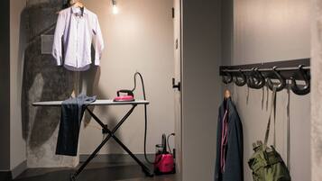A room with a hanging clothes rack, an ironing board with a shirt and a towel, and a vacuum cleaner.