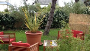 Un patio con sillas y una mesa de madera, rodeado de follaje y una gran planta en maceta.