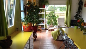 Una cocina con encimera amarilla, armarios blancos y suelo de baldosas. Hay plantas en macetas, una silla y una mesa de comedor con sillas.