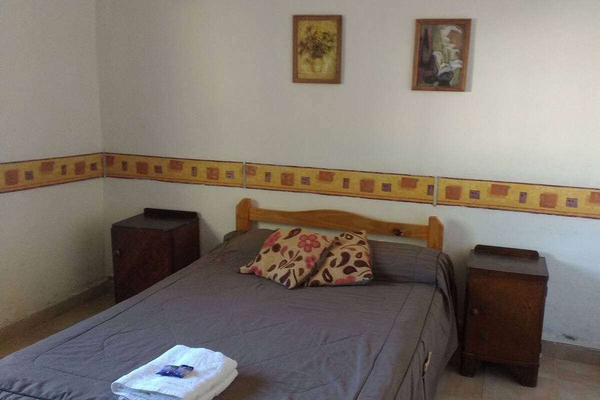 A bedroom with a bed, two nightstands, a wooden headboard, and two wall-mounted pictures.