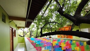 A corridor with colorful paper decorations hanging from the ceiling, a tiled floor, and a tree providing shade. - Hotel Los Ciruelos (Oaxaca)