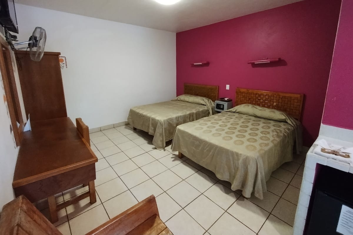 A room with two beds, a desk, a chair, a small table, and a nightstand with a lamp.